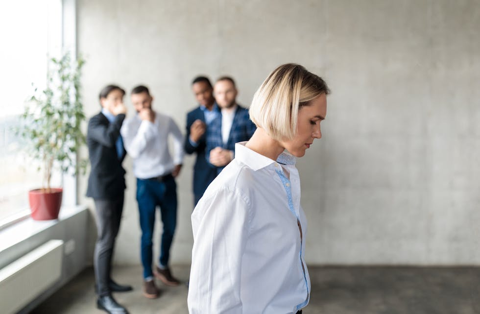 Male Colleagues Whispering Behind Back Of Unhappy Business Lady Indoors